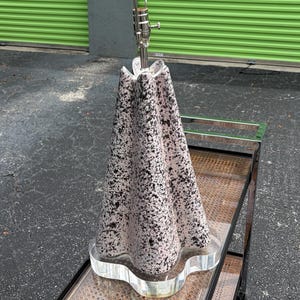 May include: A unique table lamp with a speckled, cone-shaped shade in pink and black. The lamp has a clear, wavy base and a silver-toned metal stem. The lamp is on a metal and glass side table.
