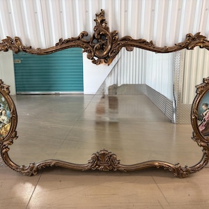 May include: Ornate, antique-style mirror with a gold-toned, scrolling frame. The mirror features two oval-shaped paintings on each side and a large central mirror. The paintings depict figures in a classical setting.
