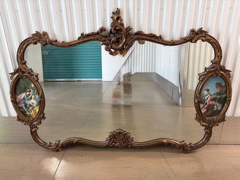 May include: Ornate, antique-style mirror with a gold-toned, scrolling frame. The mirror features two oval-shaped paintings on each side and a large central mirror. The paintings depict figures in a classical setting.