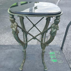 K&ouml;nnte beinhalten: Ein kleiner, quadratischer, gr&uuml;ner Metalltisch mit einer schwarzen Marmorplatte. Der Tisch hat vier Beine, die jeweils die Form einer Meerjungfrau mit einem Schwanz haben.