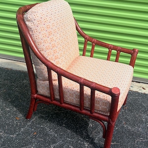 Könnte beinhalten: Ein Sessel mit einem dunkel rotbraun gebeizten Bambusrahmen und hellen Sitz- und Rückenpolstern mit einem kleinen, sich wiederholenden geometrischen Muster in Orange und Weiß. Der Sessel hat eine geschwungene Rückenlehne und vertikale Stützen.