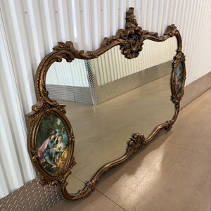 May include: Ornate gold-toned mirror with a decorative frame. The mirror features an oval painting on each side depicting a couple in a garden setting. The frame has a scrolling design and a decorative top.