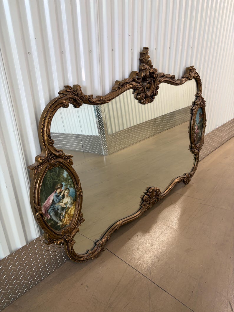 May include: Ornate gold-toned mirror with a decorative frame. The mirror features an oval painting on each side depicting a couple in a garden setting. The frame has a scrolling design and a decorative top.