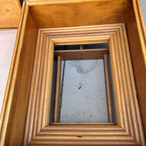 May include: A light brown, rectangular wooden frame with a layered, ridged design. The frame is open in the center and appears to be part of a larger piece of furniture, possibly a table or a decorative element.