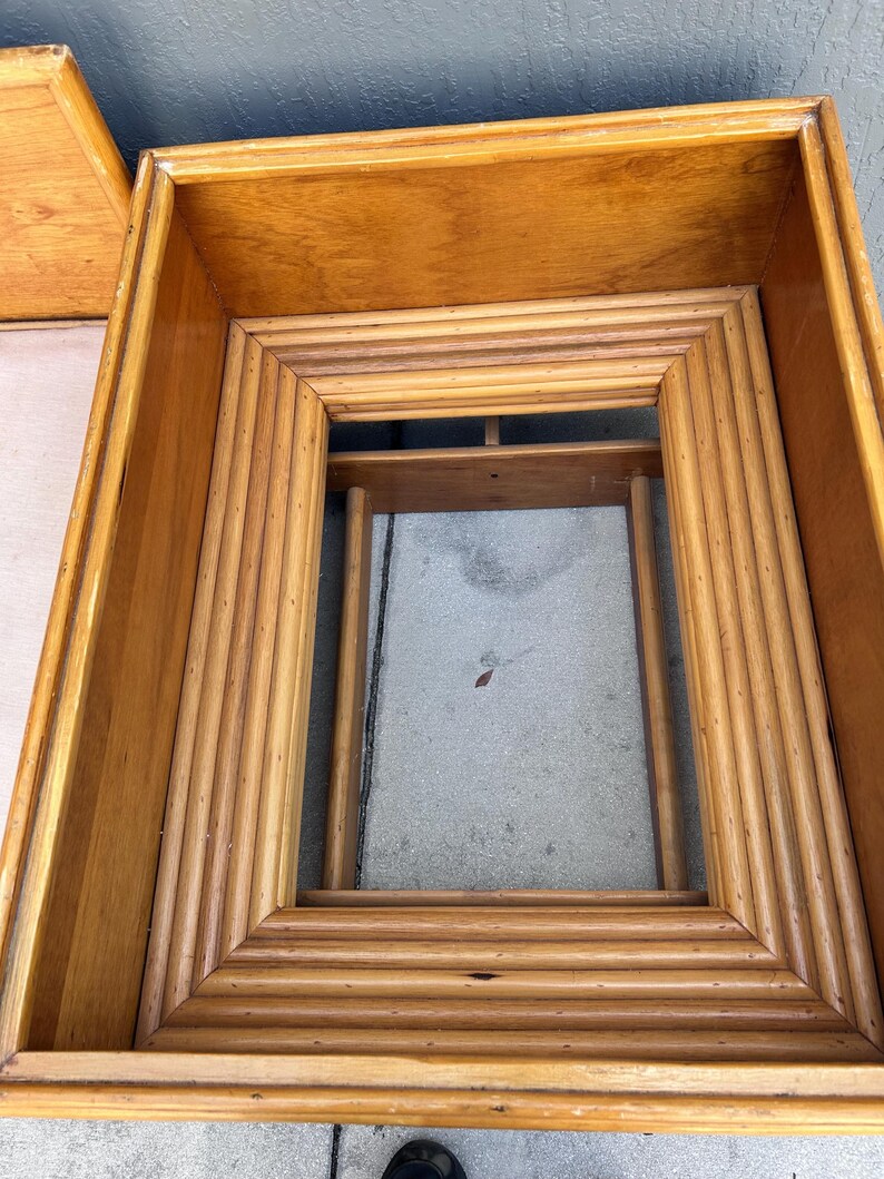 May include: A light brown, rectangular wooden frame with a layered, ridged design. The frame is open in the center and appears to be part of a larger piece of furniture, possibly a table or a decorative element.