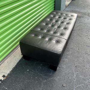 May include: A rectangular black ottoman with a tufted top and silver-colored button accents. The ottoman is made of a smooth, leather-like material and sits on short, square feet. The piece is approximately 48 inches long.