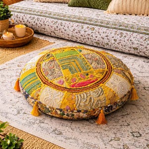 May include: A round floor cushion featuring a patchwork design of yellow, gold, and cream fabrics with intricate embroidery, floral patterns, and geometric stitching. The cushion is accented with small yellow tassels around the circular edge.