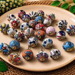 May include: A collection of round ceramic cabinet knobs featuring various hand-painted floral and geometric patterns in colours including blue, red, yellow, orange, pink, and white. Each knob has a metallic gold-toned centre screw attachment.