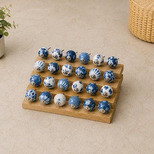 Pu&ograve; includere: Una collezione di ventiquattro pomelli rotondi in ceramica per mobili disposti su un espositore in legno a gradini. Ogni pomello presenta un motivo unico in blu e bianco, inclusi disegni floreali, a righe e geometrici.