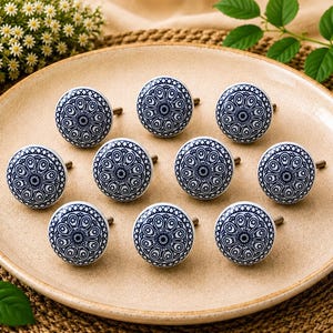 Puede incluir: Un juego de diez pomos redondos de cerámica para armarios con un intrincado patrón de estilo mandala en azul oscuro y blanco. Cada pomo tiene un acabado liso y brillante con un tornillo de metal en la parte posterior, dispuestos sobre un plato.