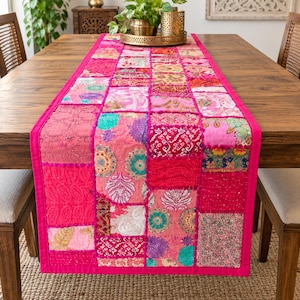 May include: A rectangular table runner featuring a patchwork design of various pink, magenta, and fuchsia fabric squares. The runner displays a mix of floral, paisley, and geometric patterns with embroidered details and a solid pink border trim.