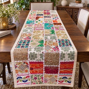 May include: A rectangular table runner featuring a patchwork design of various fabric squares in vibrant colors including red, orange, green, blue, and gold. The squares display diverse floral and geometric patterns, joined by visible white stitching.