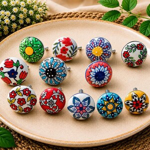 May include: A collection of eleven round ceramic cabinet knobs featuring various hand-painted floral and geometric patterns in vibrant colours including red, blue, green, and yellow, each with a small metal screw protruding from the back.