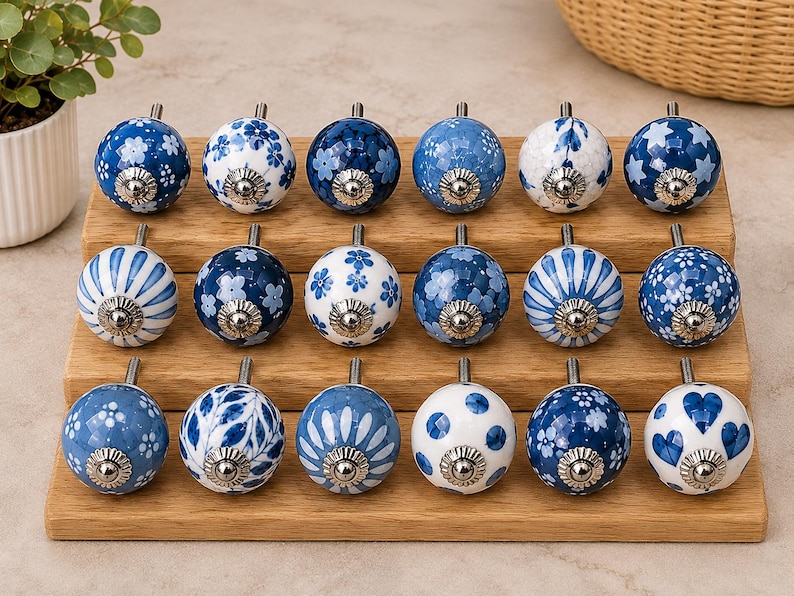 Pu&ograve; includere: Diciotto pomelli rotondi in ceramica per mobili disposti su tre file su un espositore in legno. Ogni pomello presenta un motivo unico in blu e bianco, inclusi disegni floreali, a righe, a pois, a stelle e a cuori con finiture argentate.