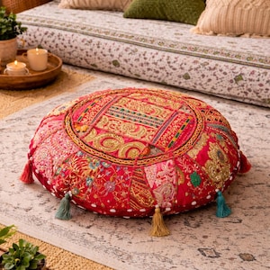 May include: A round, red floor cushion with intricate embroidery and gold accents. The cushion features colorful floral patterns and decorative tassels in various colors. It is placed on a patterned rug, suggesting a bohemian or global decor style.