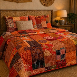 Op de afbeelding: Een patchwork sprei met een verscheidenheid aan rechthoekige en vierkante stoffen panelen in tinten oranje, rood, bruin en goud. De sprei toont diverse bloemmotieven, geometrische patronen en paisley-prints met zichtbare stiksels.