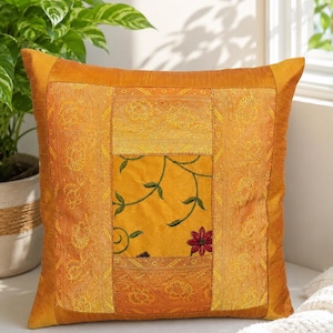 May include: A square decorative cushion featuring a patchwork design in shades of gold and mustard yellow. The centre panel displays a floral vine pattern with a single red flower, surrounded by textured borders with embroidered golden floral motifs.