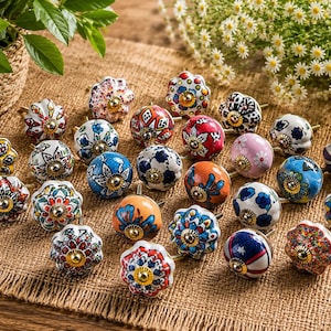 Op de afbeelding: Een collectie van vierentwintig ronde keramische kastknoppen met diverse kleurrijke handgeschilderde bloemmotieven, geometrische en abstracte patronen. Elke knop heeft een centraal goudkleurig metalen onderdeel en een geschulpte of gladde rand.