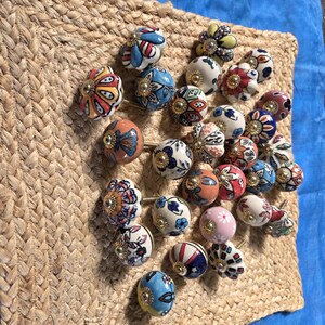 May include: A collection of round ceramic cabinet knobs featuring various hand-painted floral and geometric patterns in colours including red, blue, yellow, pink, and white, each finished with a central gold-toned metal hardware accent.