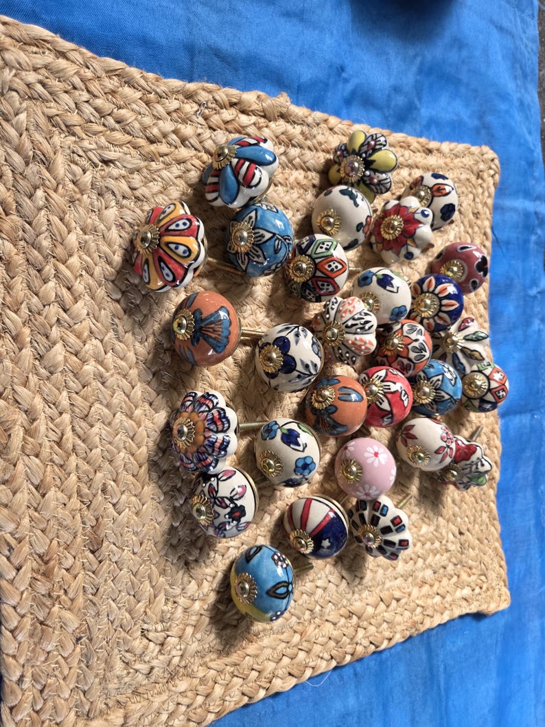 May include: A collection of round ceramic cabinet knobs featuring various hand-painted floral and geometric patterns in colours including red, blue, yellow, pink, and white, each finished with a central gold-toned metal hardware accent.