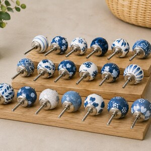 Pu&ograve; includere: Una collezione di diciotto pomelli rotondi in ceramica per mobili con vari motivi blu e bianchi, inclusi disegni floreali, a righe, a cuore e a stella. Ogni pomello ha un perno in metallo color argento ed &egrave; disposto su un espositore in legno a tre livelli.
