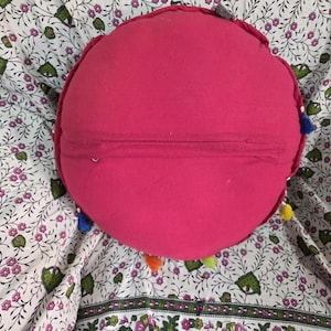 May include: A round, solid hot pink cushion featuring a horizontal zipper closure across the center. The edges of the circular pillow are decorated with small, multi-colored fabric tassels in shades of blue, yellow, orange, and green.