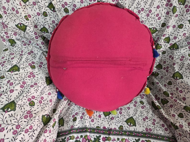 May include: A round, solid hot pink cushion featuring a horizontal zipper closure across the center. The edges of the circular pillow are decorated with small, multi-colored fabric tassels in shades of blue, yellow, orange, and green.
