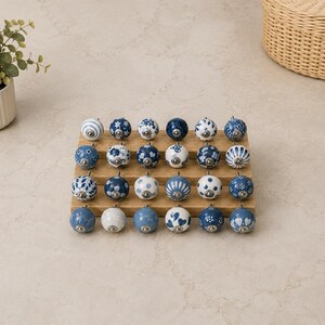 Pu&ograve; includere: Una collezione di ventiquattro pomelli rotondi in ceramica per mobili disposti in quattro file su un espositore in legno. Ogni pomello presenta un motivo unico blu e bianco, inclusi disegni floreali, a pois, a righe e a cuore con raccordi.