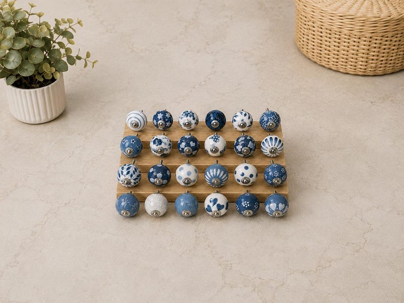 Pu&ograve; includere: Una collezione di ventiquattro pomelli rotondi in ceramica per mobili disposti in quattro file su un espositore in legno. Ogni pomello presenta un motivo unico blu e bianco, inclusi disegni floreali, a pois, a righe e a cuore con raccordi.