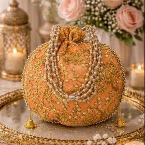 Puede incluir: Un bolso redondo de color melocotón con cierre de cordón, que presenta un intrincado bordado floral dorado y un asa de perlas. El bolso está adornado con pequeñas borlas doradas en los laterales y una delicada correa de perlas, creando un aspecto texturizado.