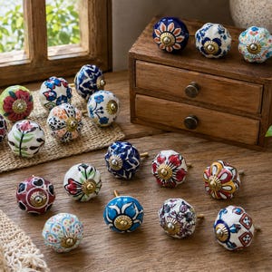 May include: A selection of decorative ceramic cabinet knobs in various colours and patterns. The knobs feature floral and geometric designs with brass hardware. They are arranged on a wooden surface near a small wooden drawer and a window.
