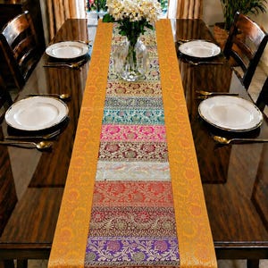 May include: A long rectangular table runner featuring a vibrant patchwork of horizontal panels in various colors including gold, teal, pink, red, and purple, all decorated with intricate floral patterns and bordered by a wide, textured gold trim.