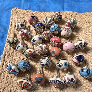 May include: A collection of round ceramic cabinet knobs featuring various hand-painted floral, geometric, and abstract patterns in colours including blue, red, orange, pink, and green, each with a central brass-toned metal mounting screw.