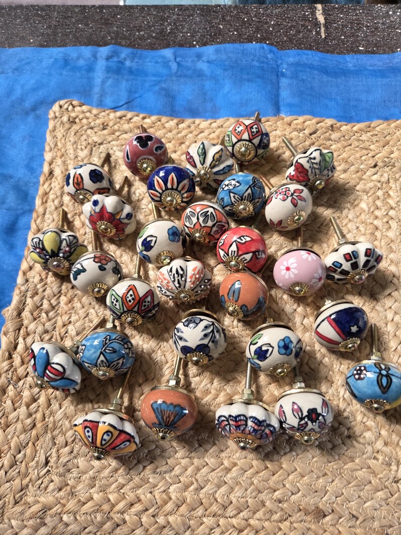 May include: A collection of round ceramic cabinet knobs featuring various hand-painted floral, geometric, and abstract patterns in colours including blue, red, orange, pink, and green, each with a central brass-toned metal mounting screw.
