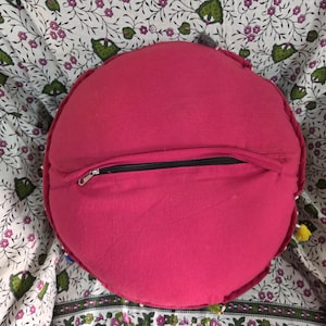 May include: A round, solid hot pink cushion featuring a black zipper closure running horizontally across the center of the back panel. The fabric appears to be a plain weave textile, and the circular shape is consistent throughout the design.