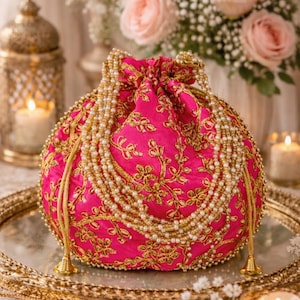 Puede incluir: Un bolso rosa intenso con cordón, adornado con bordados dorados y una cadena de perlas. El bolso redondo descansa sobre una bandeja dorada. El fondo incluye flores, velas y faroles decorativos.
