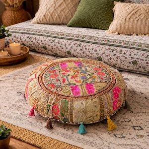 Puede incluir: Cojín redondo de suelo de patchwork con un diseño floral en rosa, amarillo y azul. El cojín tiene un borde dorado y está adornado con borlas de colores. El cojín está colocado sobre una alfombra estampada, con cojines y un sofá floral en el fondo.