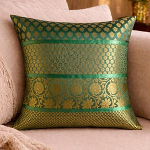 Könnte beinhalten: Dekoratives grünes und goldenes Kissen mit floralem Muster. Das quadratische Kissen besteht aus glänzendem Stoff. Das Design umfasst goldene Blumenmuster und grüne Bänder. Das Kissen liegt auf einem hellrosa Sofa.