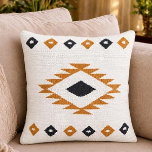 May include: Square white throw pillow with a geometric design in black and gold. The pillow features a central diamond shape with a series of smaller diamonds above and below. The pillow is placed on a beige sofa.