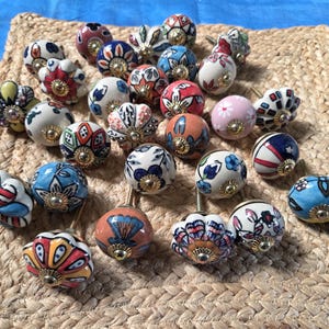 May include: A collection of round ceramic cabinet knobs featuring various colourful hand-painted floral and geometric patterns. Each knob has a central gold-toned metal hardware piece and a smooth, glossy finish, showcasing a diverse array of designs.