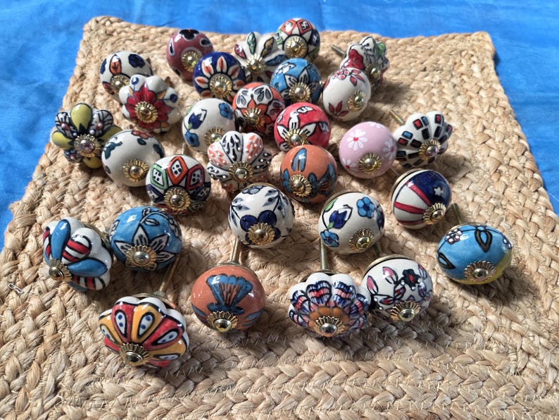 May include: A collection of round ceramic cabinet knobs featuring various colourful hand-painted floral and geometric patterns. Each knob has a central gold-toned metal hardware piece and a smooth, glossy finish, showcasing a diverse array of designs.