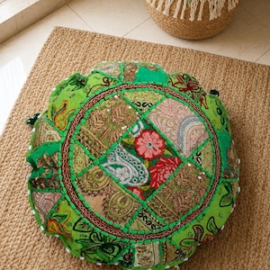 May include: A round floor cushion featuring a vibrant green fabric base with a patchwork design. The top surface is decorated with intricate embroidery, metallic gold thread accents, small reflective sequins, and floral patterns in red and pink.
