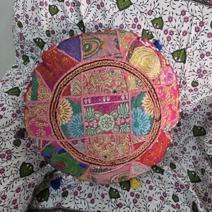 May include: A round floor cushion featuring a patchwork design of pink, gold, blue, and green fabric panels. The surface is decorated with intricate embroidery, metallic thread stitching, and small reflective mirror accents arranged in a circular pattern.