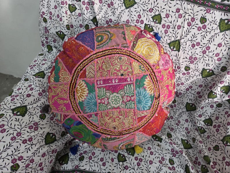 May include: A round floor cushion featuring a patchwork design of pink, gold, blue, and green fabric panels. The surface is decorated with intricate embroidery, metallic thread stitching, and small reflective mirror accents arranged in a circular pattern.
