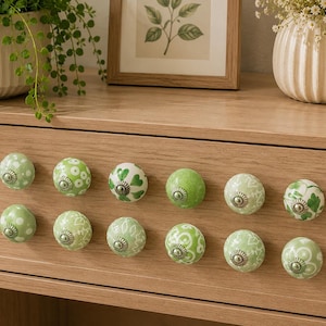 Puede incluir: Un cajón de madera con doce pomos decorativos de cerámica en tonos verdes y blancos. Los pomos presentan varios diseños florales y de puntos. El cajón forma parte de un mueble de madera clara.