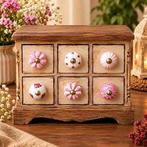 May include: A wooden storage chest with six drawers, each featuring a unique ceramic knob. The chest has a distressed finish, and the drawers are painted in an off-white color. The knobs vary in shape and pattern, including pink floral, red polka dot, and solid pink designs with brass-toned centers.