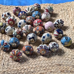 May include: A collection of round ceramic cabinet knobs featuring various colourful hand-painted floral and geometric patterns. Each knob has a central gold-toned metal hardware piece and a smooth, glossy finish with diverse designs in red, blue, pink, and white.