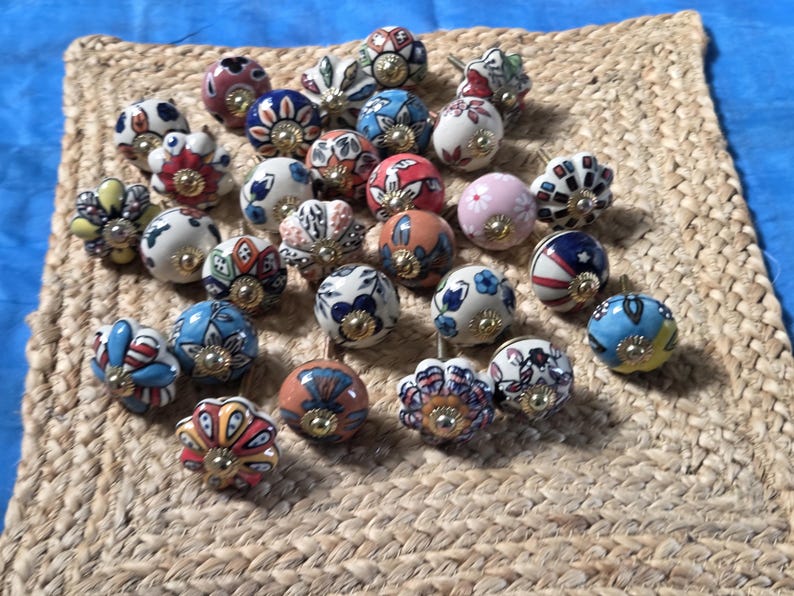 May include: A collection of round ceramic cabinet knobs featuring various colourful hand-painted floral and geometric patterns. Each knob has a central gold-toned metal hardware piece and a smooth, glossy finish with diverse designs in red, blue, pink, and white.