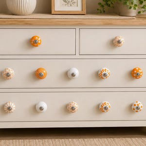 May include: A cream-colored wooden dresser with a light wood top, featuring two small top drawers and two wide bottom drawers. The drawers are fitted with twelve round ceramic knobs in various orange, white, and tan patterns with silver hardware.