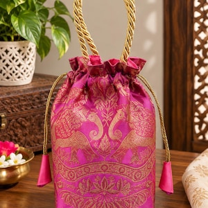 May include: A vibrant fuchsia drawstring pouch featuring intricate gold-toned woven patterns of elephants and floral motifs. The bag includes a twisted gold rope handle and matching drawstring cords with fuchsia fabric tassels at the ends.